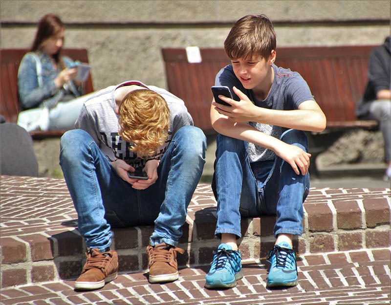 two boys sitting next to each other one is looking down and the other is looking at his phone